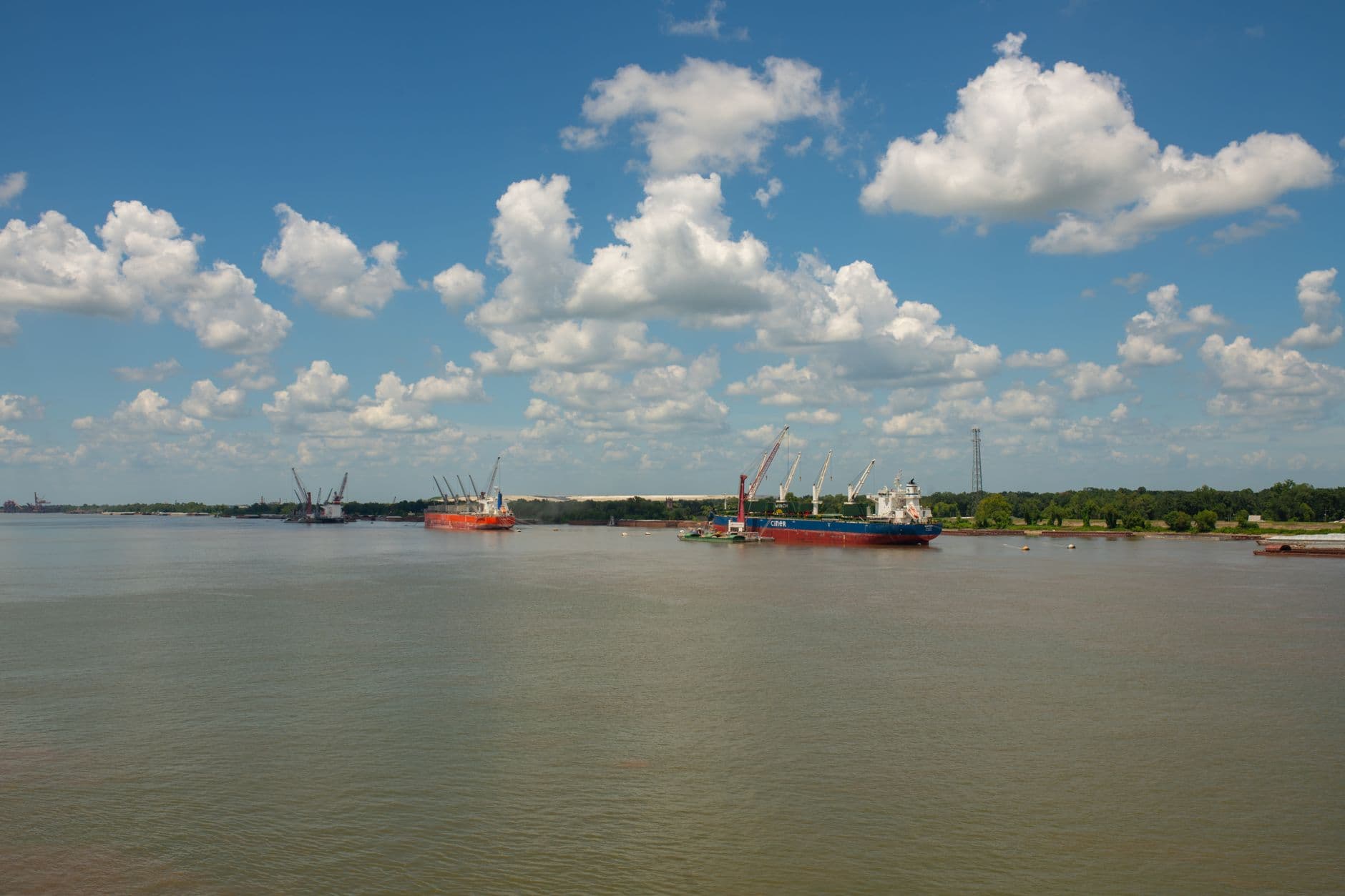 Cargo ships anchored on a calm river with a clear blue sky and fluffy clouds.